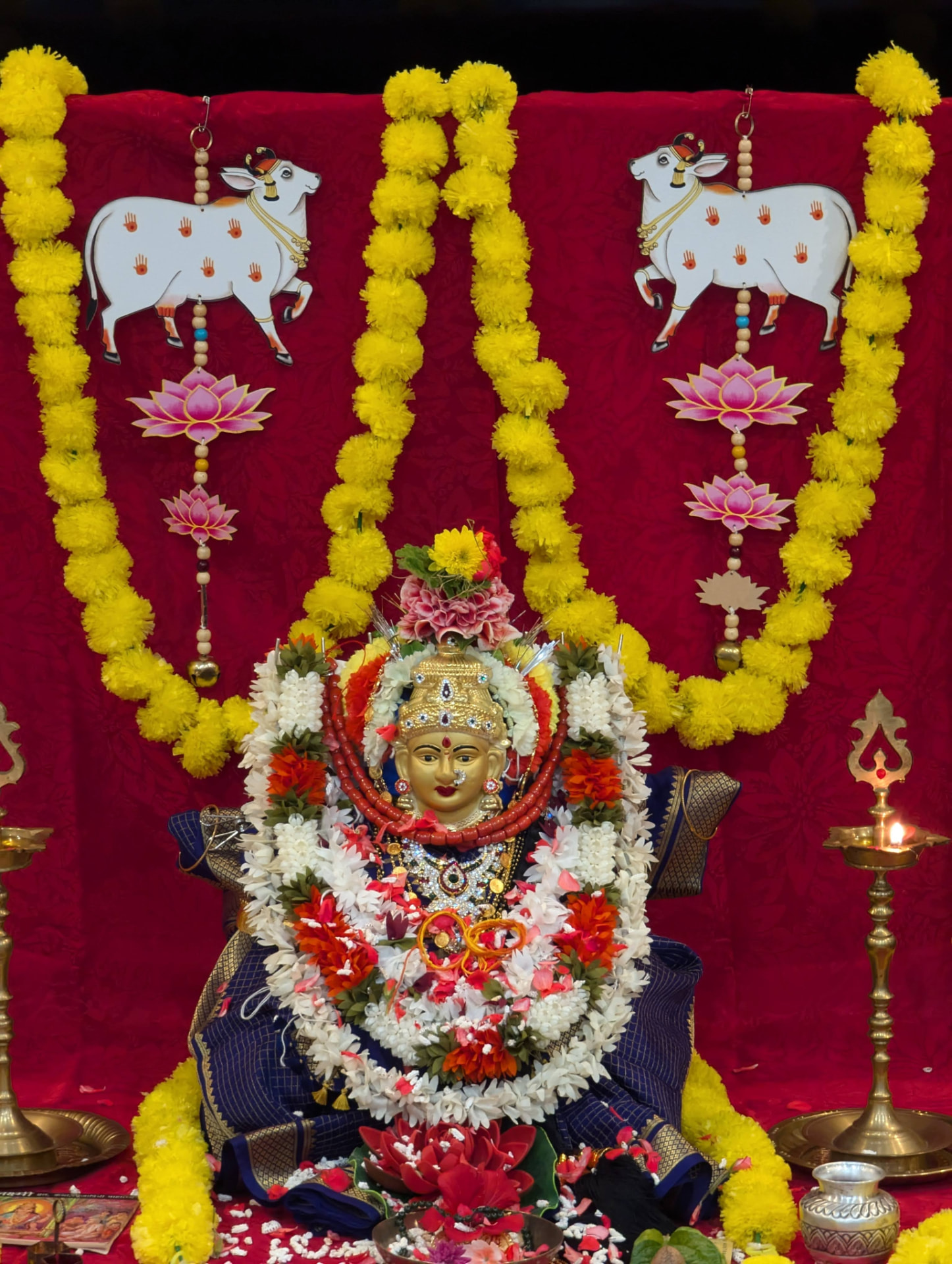 Varamahalakshmi Devi adorned in silk, coral and jasmine, with Kamadhenu and lotus torans on the crimson backdrop